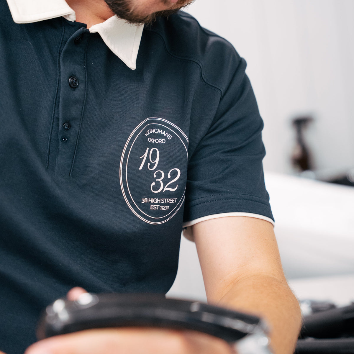 A close-up lifestyle shot of a man wearing a dark navy polo shirt with a white collar and a circular white graphic on the chest. He is holding a black object in his hands, with a blurred indoor background.