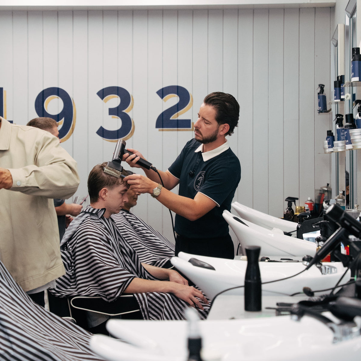 A barbershop interior with a barber styling a customer's hair. The barber wears a dark navy polo shirt, while the customer sits in a barber chair wearing a black-and-white striped cape.